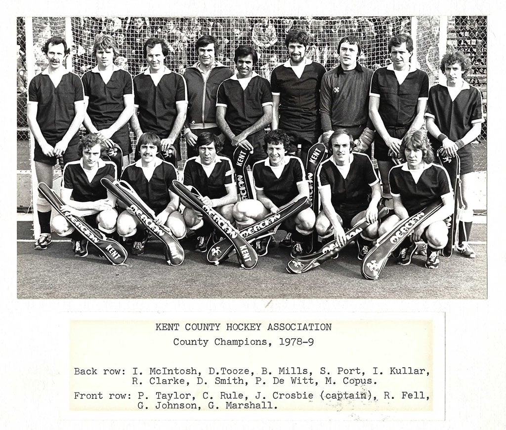 Two rows of men pose for a team photograph on a grass hockey pitch in front of the goal. They are wearing dark shirts and holding either hockey sticks or hockey stick bags with the logo of manufacturer Mercian Sports and their team Kent.