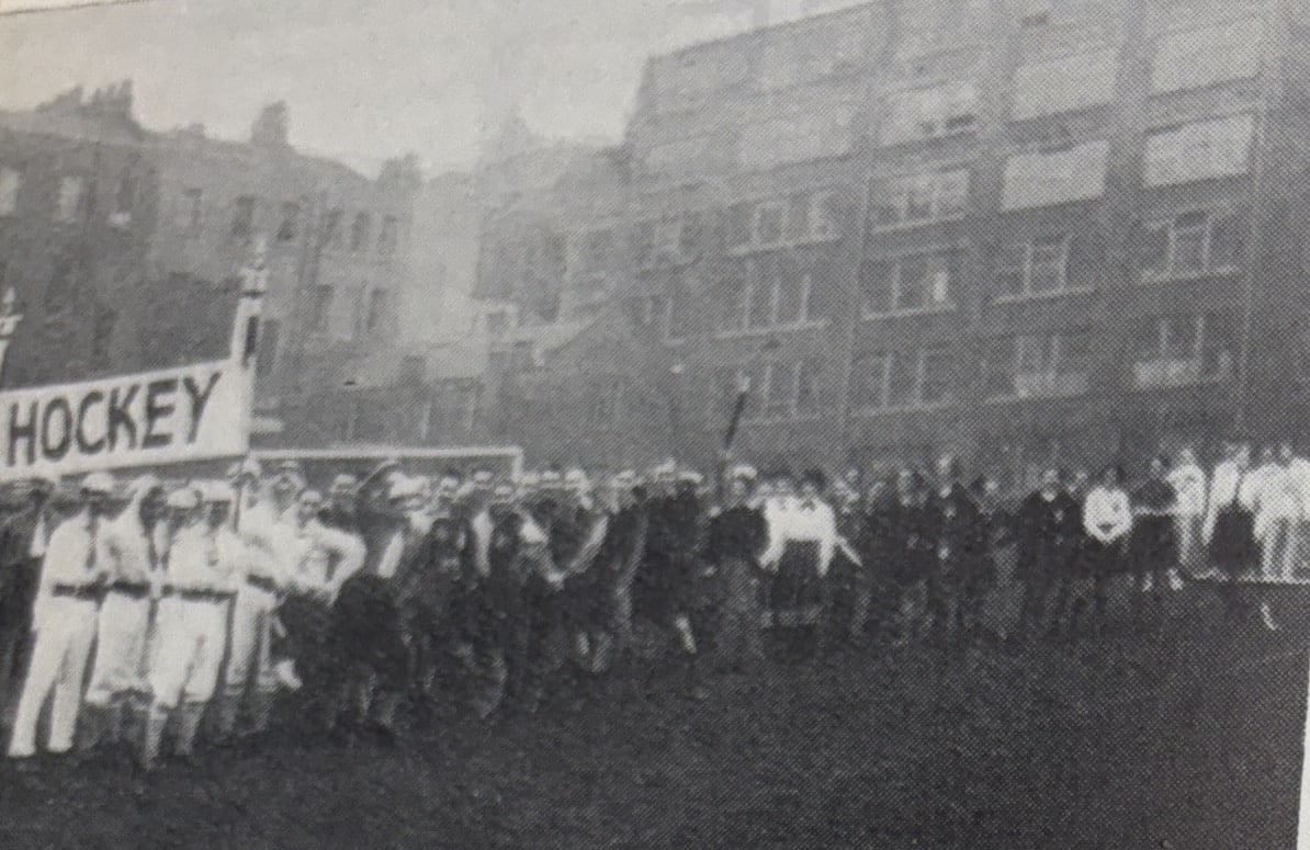 A black and white photograph depicting a column of men and women in old-style hockey clothing. They wave and cheer at the camera. At the front of the column is a large white banner on two poles with the word "HOCKEY". The backdrop is a nondescript block of flats.