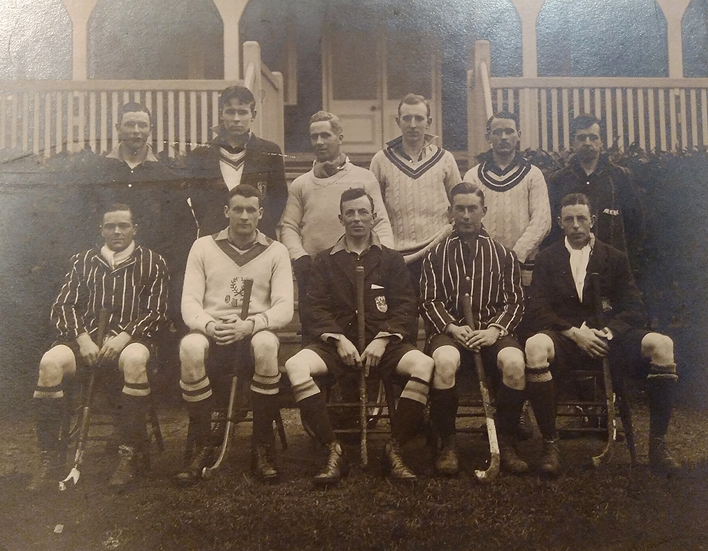 Black and white team photograph of 11 young men in two rows, the front row seated. They are wearing combination of blazers and jumpers with the insignia of their club or representative teams. There is a wooden pavilion in the background.