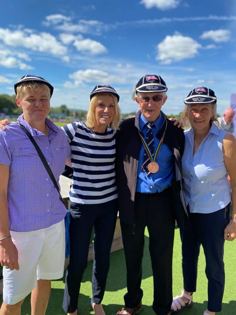 Three Caucasian women stand with an elderly Caucasian man, all smiling with a bright blue sky above them dotted with small clouds. All four people wear navy blue caps with silver trim and the Union Flag of Great Britain.