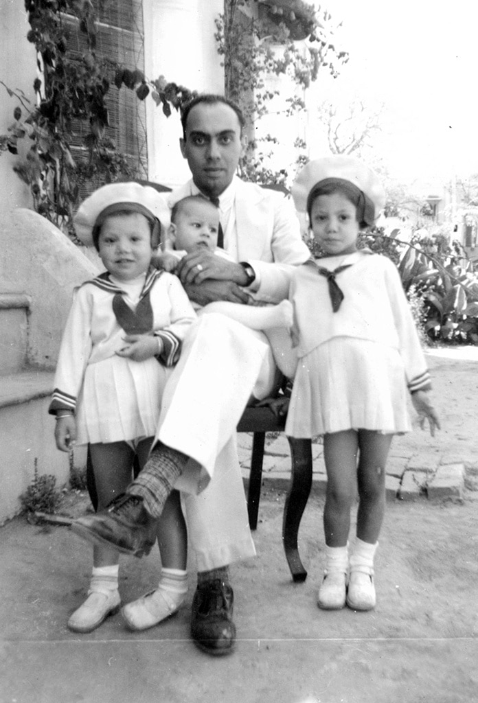 Black and white photograph of a family posed outdoors, with an adult seated and holding a child, flanked by two children in sailor outfits. The scene suggests a calm and affectionate family moment, with gentle smiles reflecting warmth and togetherness.
