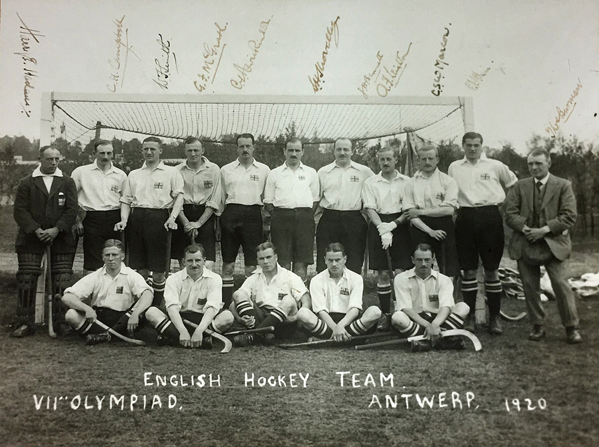 Black and white photograph of a men's hockey team positioned in two rows. They wear white shirts, each with a union flag pocket badge and dark, knee-length shorts. The players look confident and relaxed.