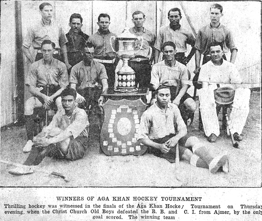 George Marthins (sitting front row, right) with the Christ Church Old Boys hockey team, winners of the Aga Khan tournament. From the George Eric Marthins scrapbook held in The Hockey Museum collection. Black and white photograph of a men's hockey team. The players stand or are seated around two trophies; a large chalice cup sits centrally on a table and a giant shield is propped against the table legs. George Marthins is positioned prominently in the front row. His attire and posture reflect the athletic spirit of the era and his dedication to the sport.