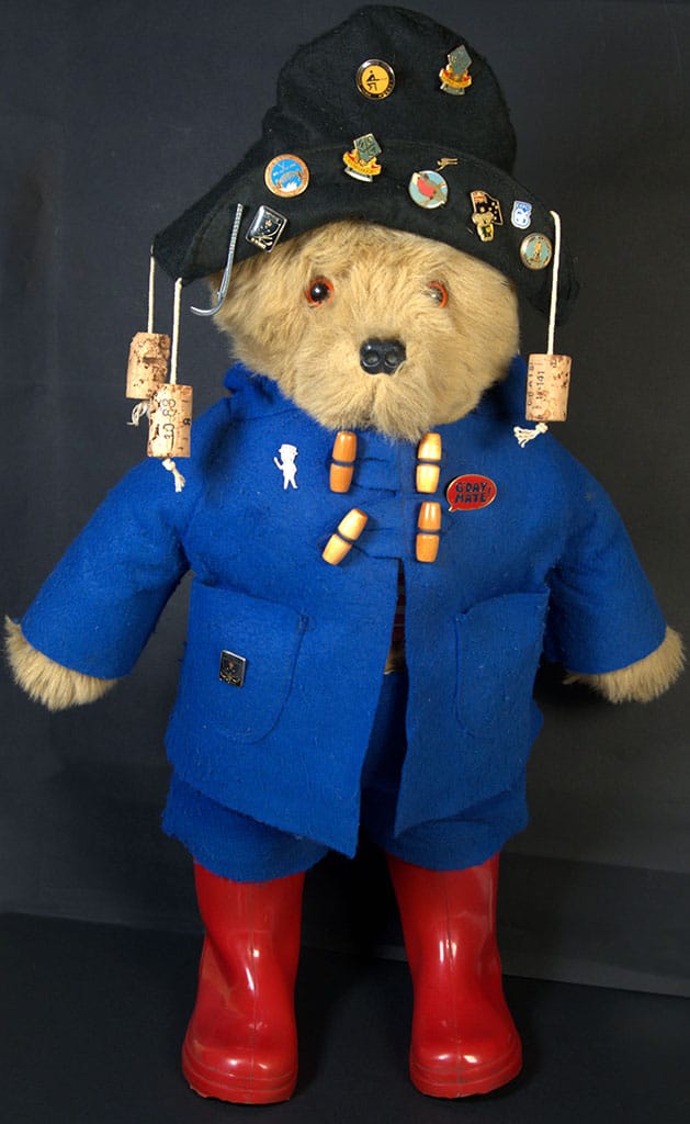 A Paddington Bear figurine dressed in a blue coat, red wellington boots, and a black hat adorned with various pin badges, displaying a cheerful expression.