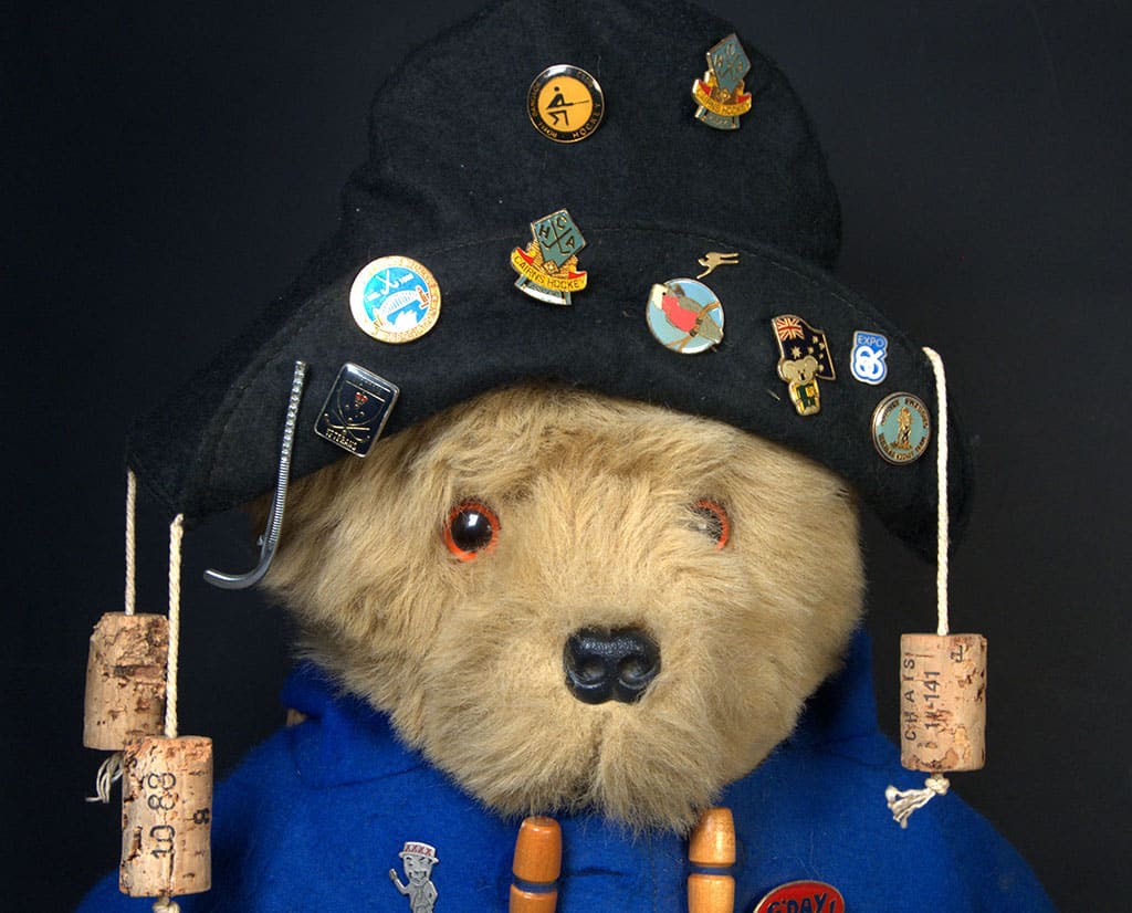 A Paddington Bear figurine wearing a blue coat with wooden toggles, a black hat covered in hockey pin badges, and cork decorations, displaying a curious expression.
