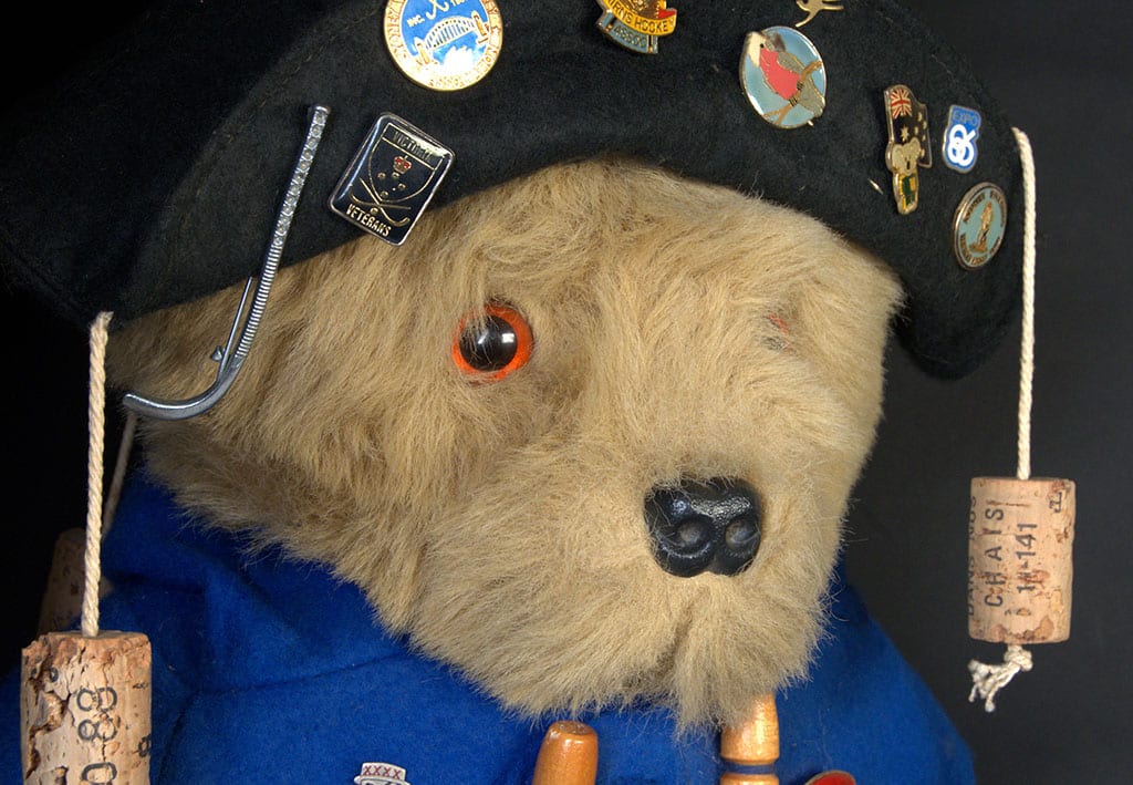 A Paddington Bear figurine adorned with a blue coat, a black hat decorated with hockey pins, and a cork accessory, showing a gentle and curious expression.