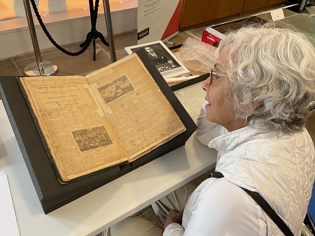 An elderly person examines an open, aged scrapbook on a table in a museum. The scrapbook displays yellowed pages with vintage text and photos, evoking a sense of curiosity and nostalgia. The scene is framed by a velvet rope barrier and informational displays in the background.