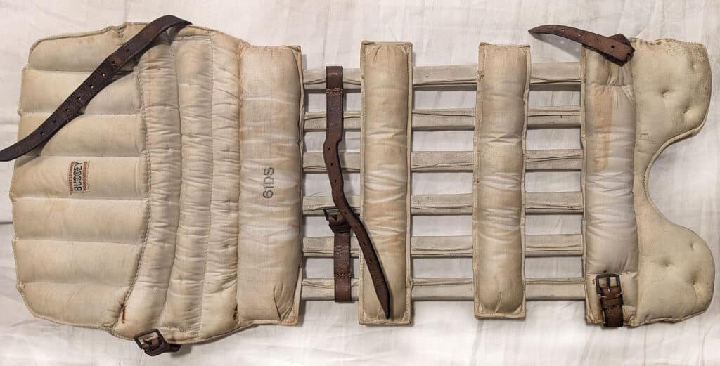 Vintage Bussey leg guard backside, featuring worn beige padded sections with horizontal cane supports, secured with brown leather straps and buckles, and marked with "61DS" and "SPORTS & GAMES MANUFACTURERS" branding.