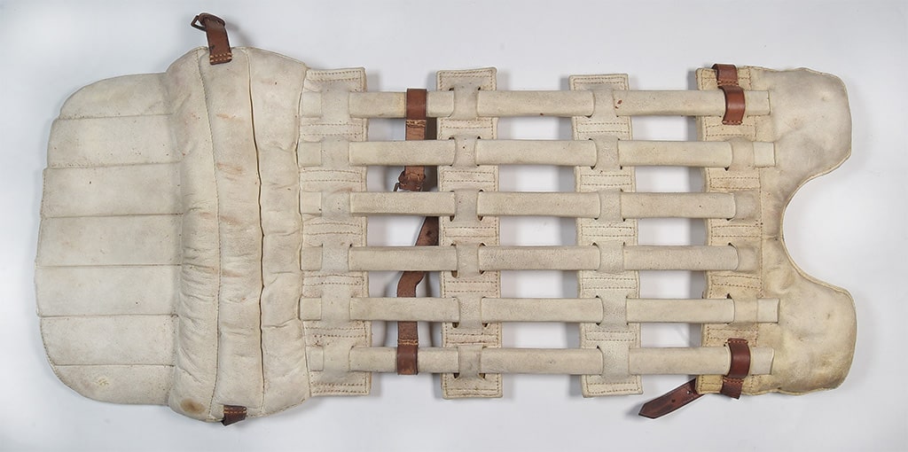 Vintage hockey pad with worn beige leather covering, featuring a padded inner section and a grid of vertical and horizontal cane supports, secured with brown leather straps and buckles.