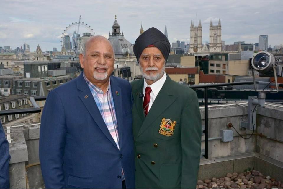 Two men standing together on a rooftop with a city skyline featuring the London Eye and Westminster Abbey in the background. The man on the left wears a blue suit with a checkered shirt, while the man on the right is dressed in a green blazer with a crest, red tie, and a turban, suggesting a formal or celebratory event related to hockey.