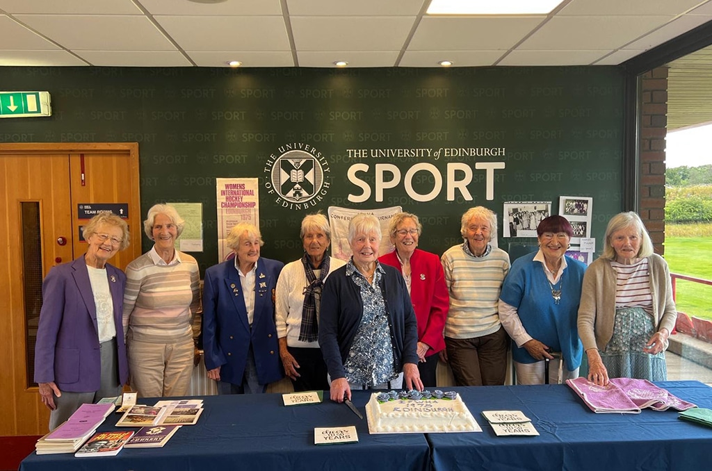 A group of elderly women standing together in front of a dark green "The University of Edinburgh Sport" backdrop, celebrating an event with a cake that reads "100 Years Edinburgh." They are dressed in colorful outfits, with some wearing blazers adorned with pins. A table in front of them holds the cake, books, pamphlets, a pink cloth, and "100 Years" signs.