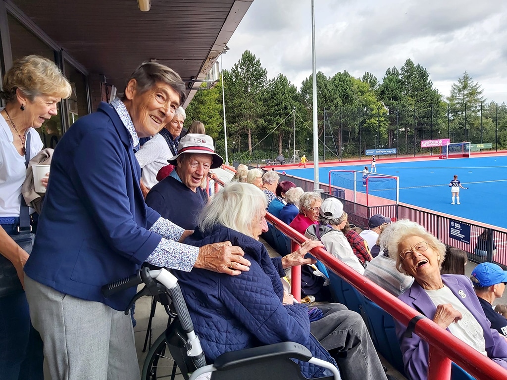 A group of elderly spectators, including some in wheelchairs, enjoying a hockey match at an outdoor stadium. One person stands behind a wheelchair, resting a hand on the occupant, while others sit on blue seats along a red railing. The blue hockey field and players are visible in the background, with trees and a cloudy sky completing the scene.