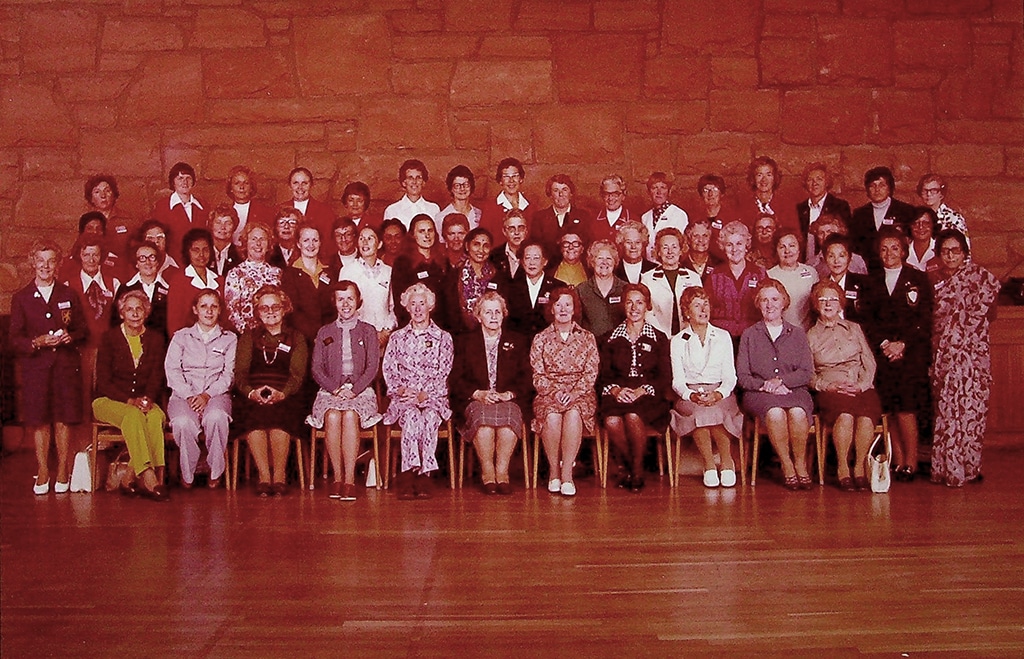 A large group of people, mostly women, pose for a group photo in front of a stone wall, dressed in a variety of colorful vintage outfits, conveying a sense of community and historical significance.
