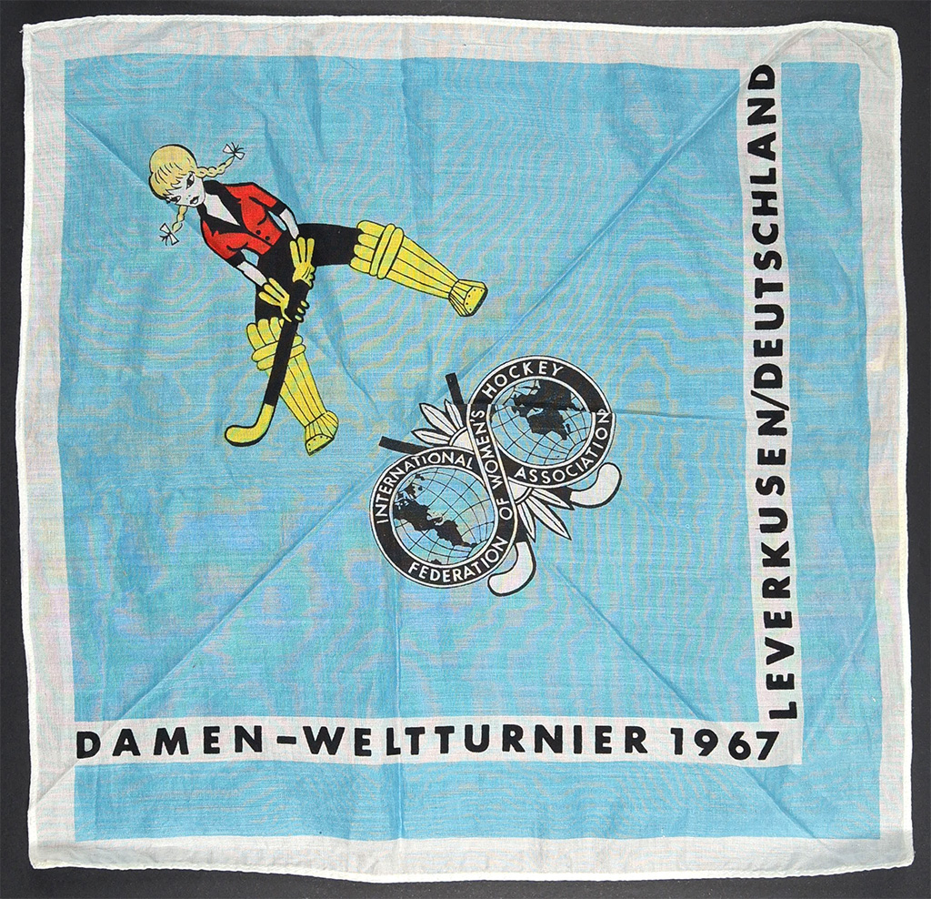 A light blue fabric handkerchief with a white border, featuring a stylized illustration of a female hockey goalkeeper in a red top, black shorts, and yellow leg guards, mid-action with a stick. A circular emblem with a globe and text is centrally placed, while black text along the edges.