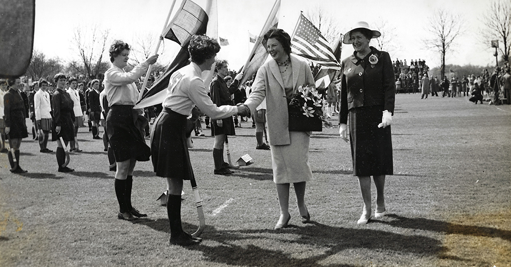 The black-and-white image shows three women in the foreground on a grassy field. Two younger women, dressed in a hockey kit of shirt, skirt, knee-high socks, and holding flags, shake hands with an older woman in a suit and hat, holding flowers. More people in similar uniforms stand in the background, some with flags, against a sparse, tree-lined horizon. The scene evokes a sense of ceremony and camaraderie.