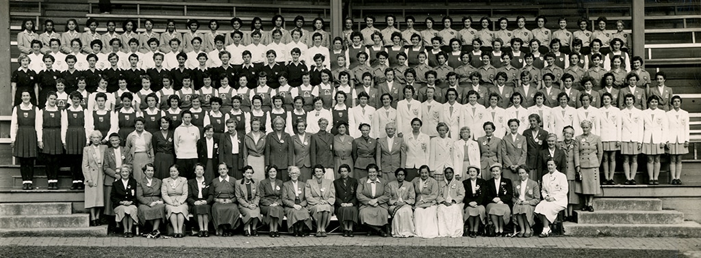 A black and white photo showing a large group of young women wearing hockey kits and, at the front, older women in formal skirts and jackets. They stand and sit in rows on concrete steps, with a stadium structure in the background, conveying a sense of unity and pride.