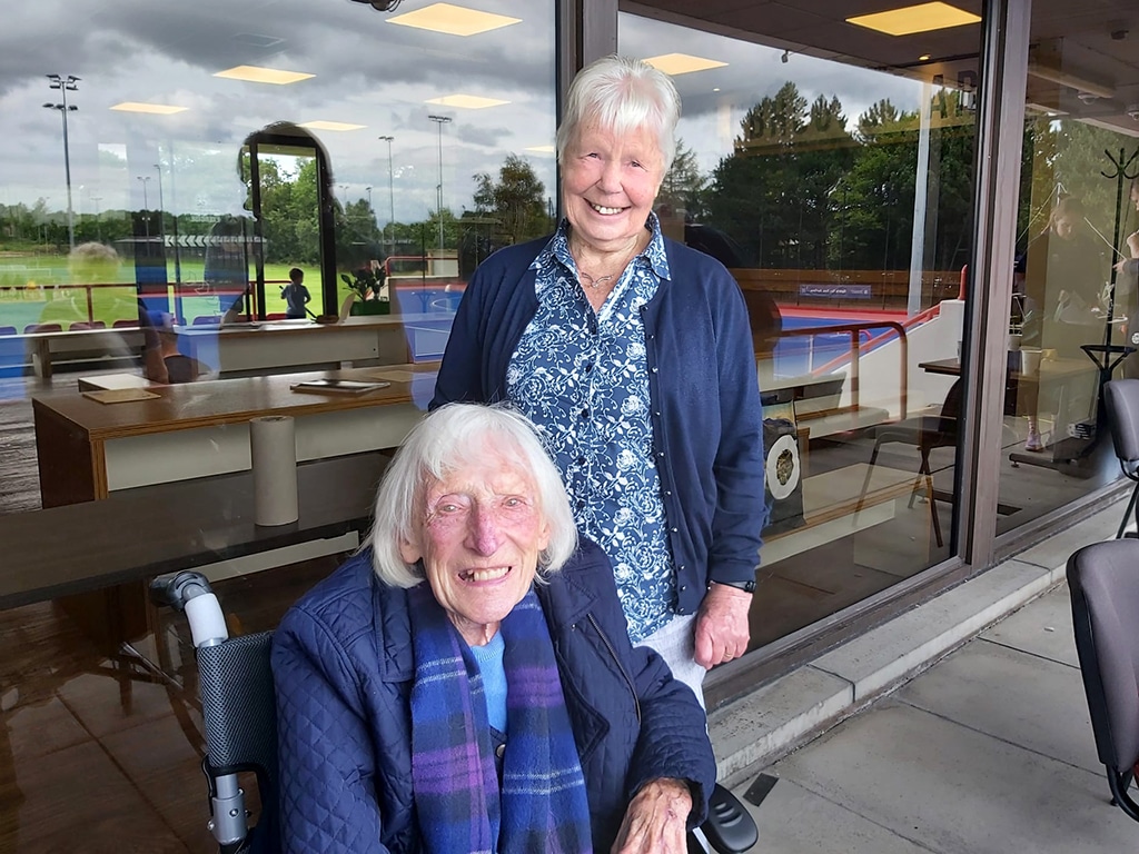 Two elderly women, one standing and one seated in a wheelchair, smiling outside a modern building with large windows. The standing woman wears a floral blouse and blue cardigan, while the seated woman is dressed in a blue jacket and colorful scarf. The background features a sports field with a blue hockey pitch reflected in the glass.