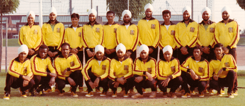 A vintage team photo of a hockey team, featuring 17 players wearing matching yellow uniforms with red and black accents. The players, some wearing turbans, are arranged in two rows with the front row kneeling on a grassy field and the back row standing, with a netted fence and track visible in the background.