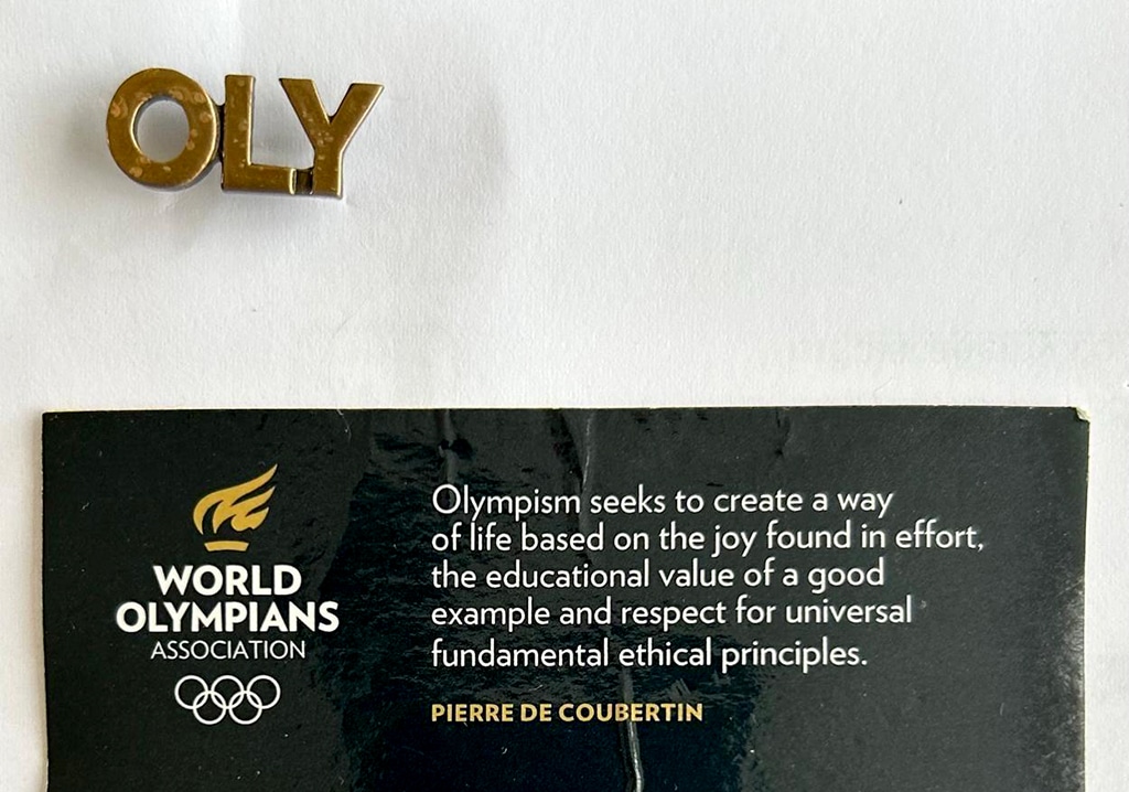 A pin badge formed of the letters "OLY" placed above a black card from the World Olympians Association. The card features the association's logo with Olympic rings and a flame, along with a quote from Olympic founder Pierre de Coubertin: "Olympism seeks to create a way of life based on the joy found in effort, the educational value of a good example and respect for universal fundamental ethical principles."