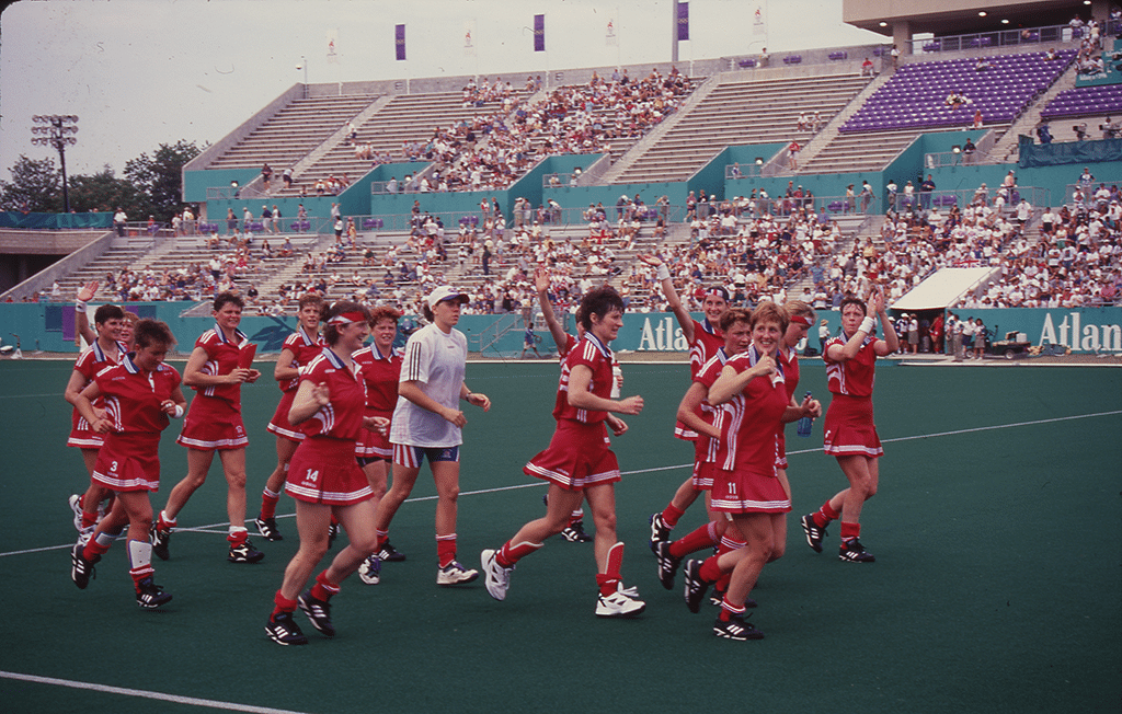 Team in red uniforms celebrates on field, waving to cheering crowd in stadium. Background shows "Atlanta" sign, purple seats. Evokes joy and triumph.