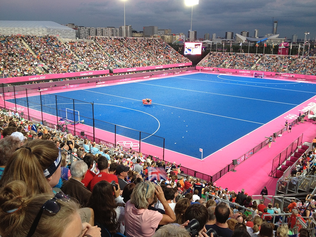 A crowded stadium with excited fans watching a hockey match on a blue pitch with a bright pink edge to the playing surface. Urban backdrop and floodlights. Evokes excitement, unity, and anticipation.