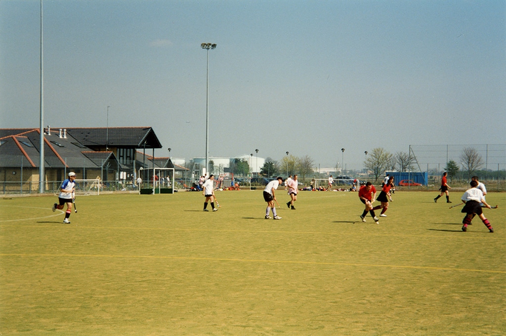Players in colourful uniforms play hockey on a sandy, green field, with buildings and trees in the background. Evokes energy, teamwork, and outdoor fun.