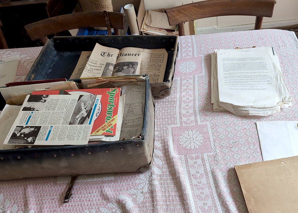 Worn boxes filled with old newspapers and documents on a pink patterned cloth. Cluttered, nostalgic feel. Evokes curiosity and history.