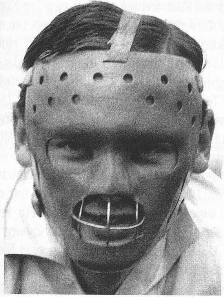 A close-up photograph of a man in a large face-mask with wide eye holes and a single mouth hole covered with a large metal grill.