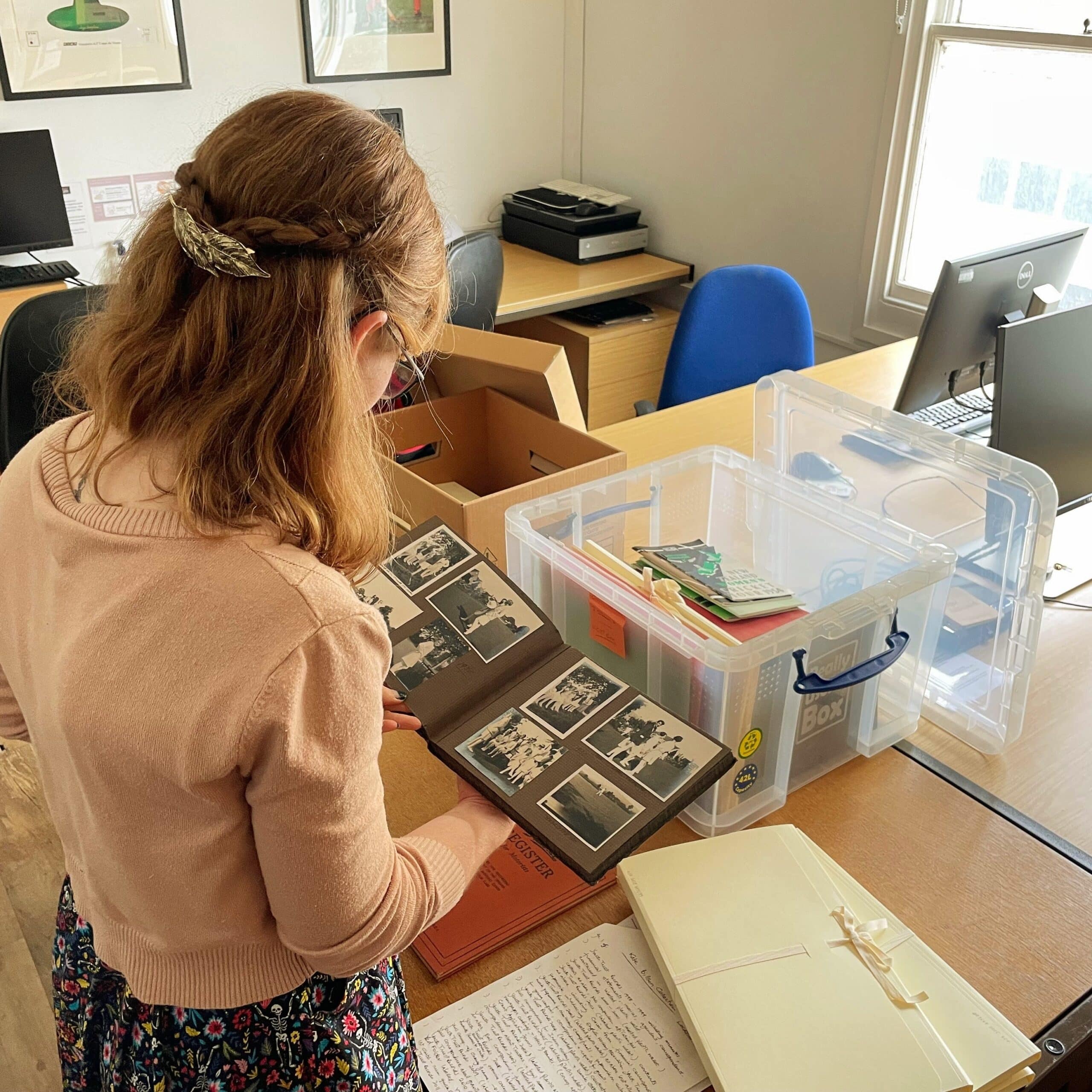 A young woman looks through boxes and packets of archival documents in a well-lit room.