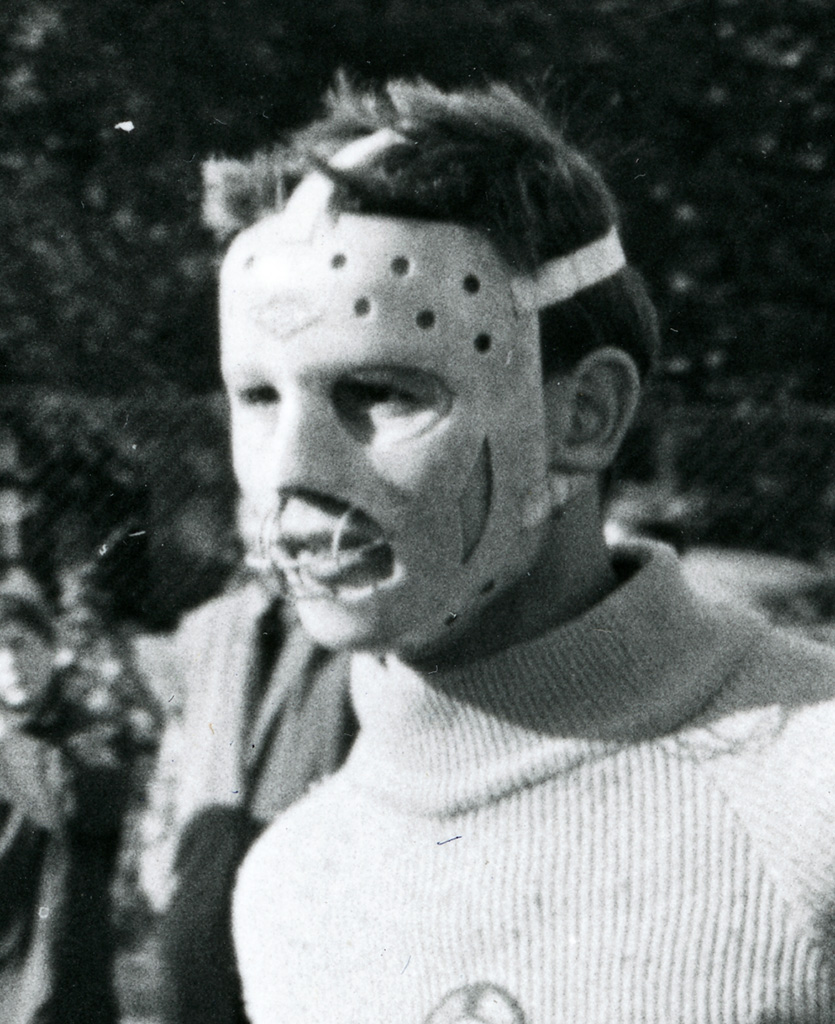 Close up photograph of a hockey goalkeeper wearing a woolly turtle-neck jumper. His face is covered by a scary-looking mask with a metal grill over the mouth.