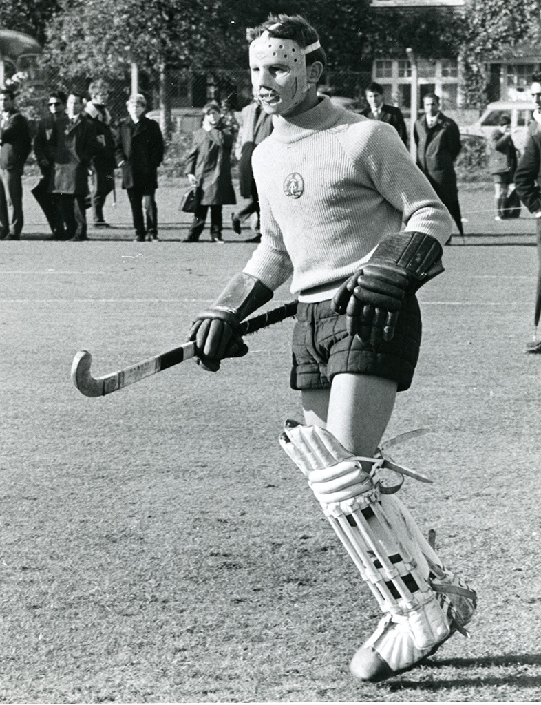 A hockey goalkeeper wearing cane leather leg guards and padded boots, padded shorts, a woolly jumper and large, heavy duty gloves. His face is covered by a scary mask with a metal grill over the mouth. Several people watch on in the background at the edge of a grass pitch.