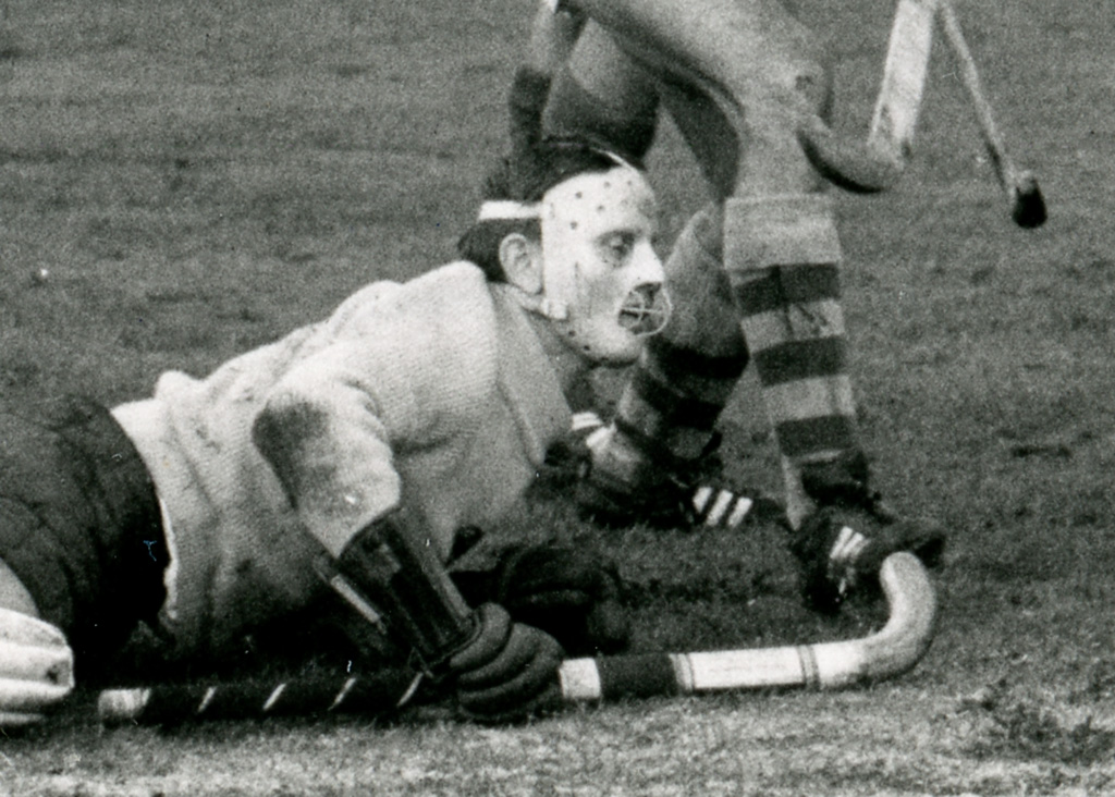 Action photograph of a hockey goalkeeper immediately after making a save. He is wearing a woolly turtle-neck jumper and large gloves lying on a grass hockey pitch. His face is covered by a scary-looking mask with a metal grill over the mouth. He is lifting himself up using his right arm which holds a hockey stick.