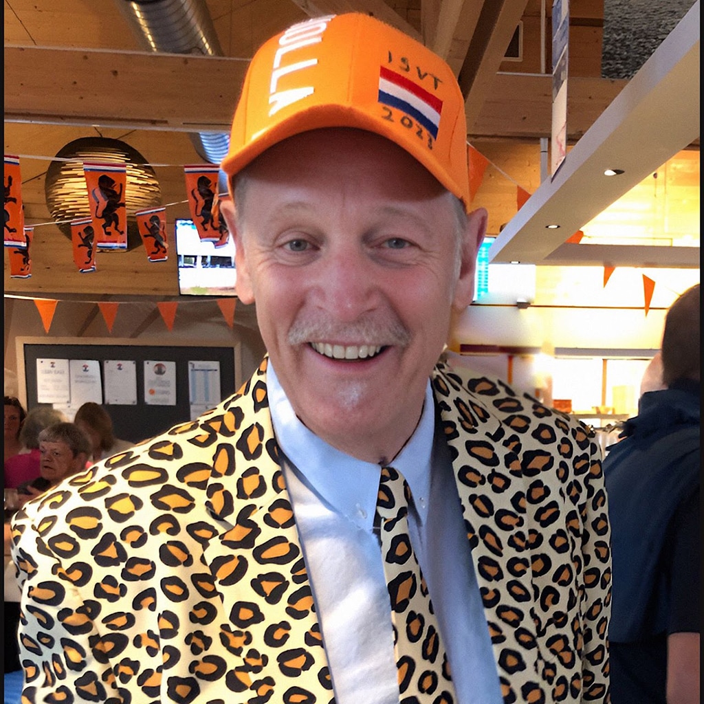A head-and-shoulders photograph of a smiling middle-aged man with a goatee bears wearing a leopard print suit and tie and an orange baseball cap