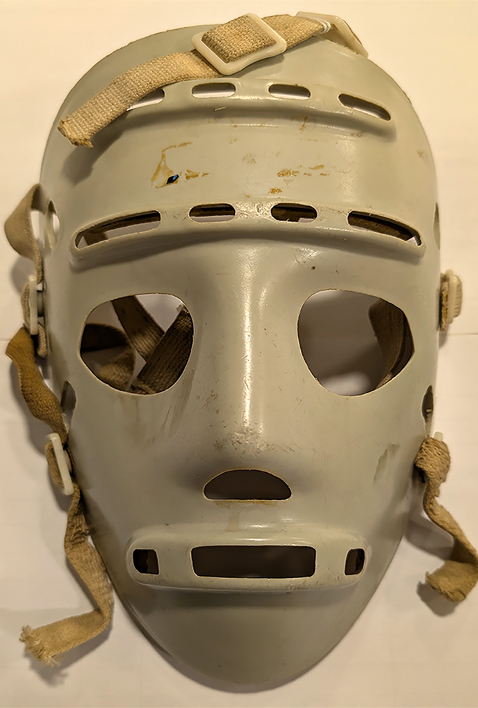 A long, thin, grey, sinister-looking plastic mask with large holes for eyes, a smaller hole for the nose and mouth shape of three long narrow slits. There are elasticated cloth straps and buckles behind.