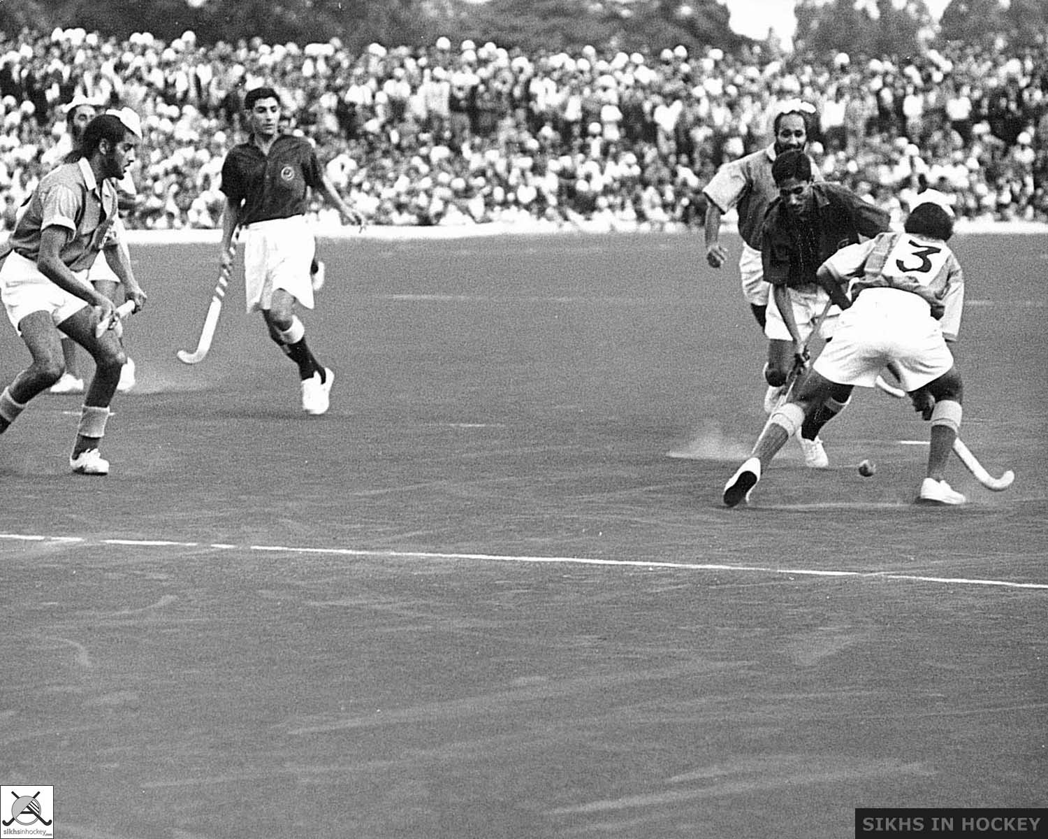 Black and white photograph of two South Asian heritage hockey teams competing on a flat dusty surface. A large crowd is in the background.