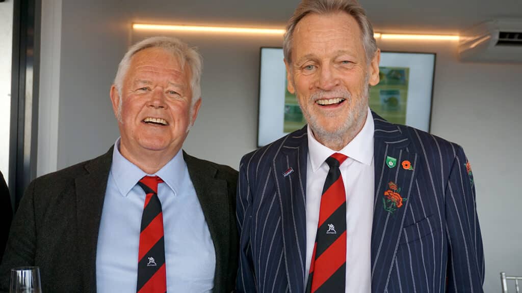 Two smiling middle-aged men wearing dark suits and matching red and black Southgate Hockey Club ties.