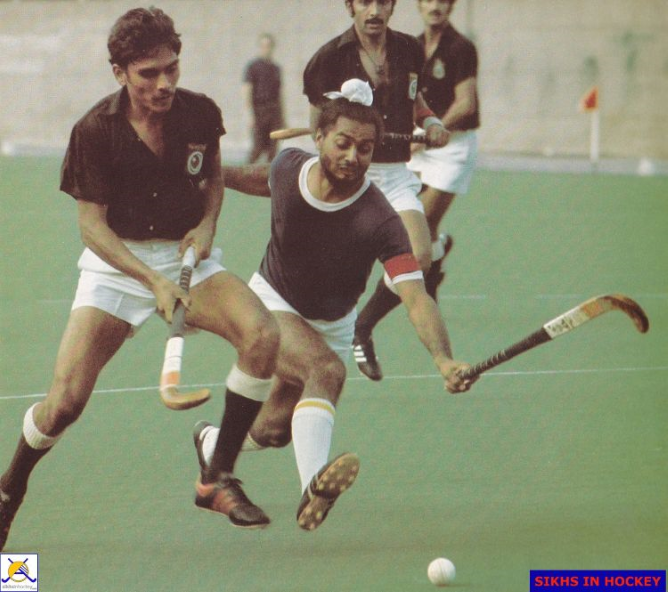 Action shot of two men of South Asian heritage competing for the ball on a flat green pitch, one dribbling the other tackling.