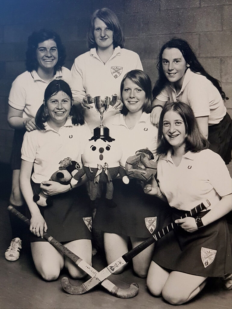 Black and white photograph of six smiling young women in polo shirts and skirts, either standing or kneeling, holding hockey sticks, toy mascots and a small trophy.