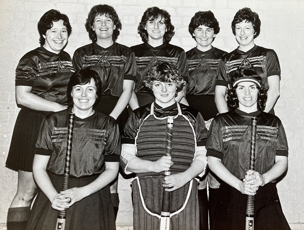 Black and white group photograph of eight smiling young women pose in front of a white-washed brick wall.