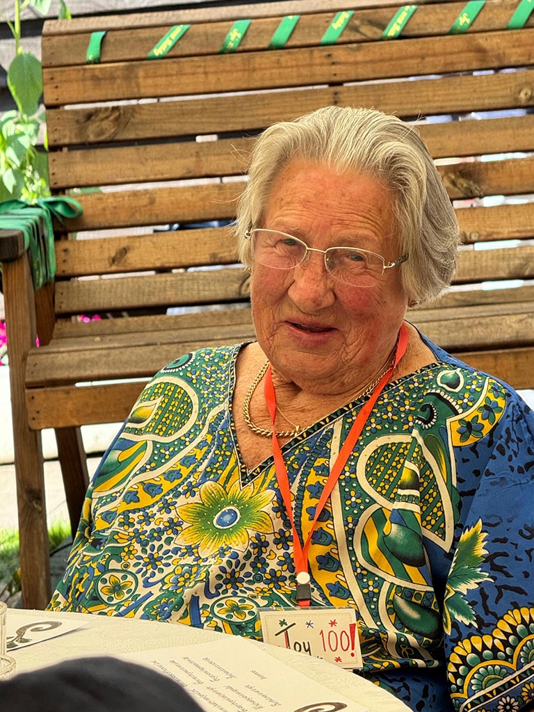 An elderly lady wearing a ornate blouse is seated at a table smiling.