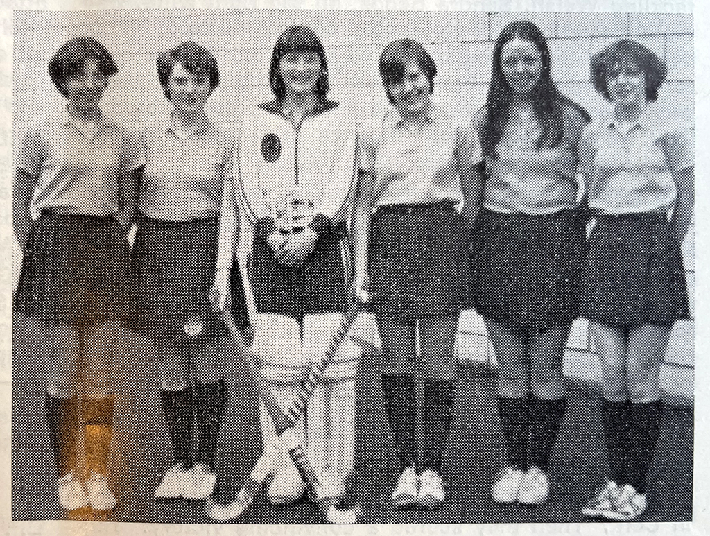 Black and white photograph of six smiling school girls in polo shirts and skirts pose in a line in front of a wall.