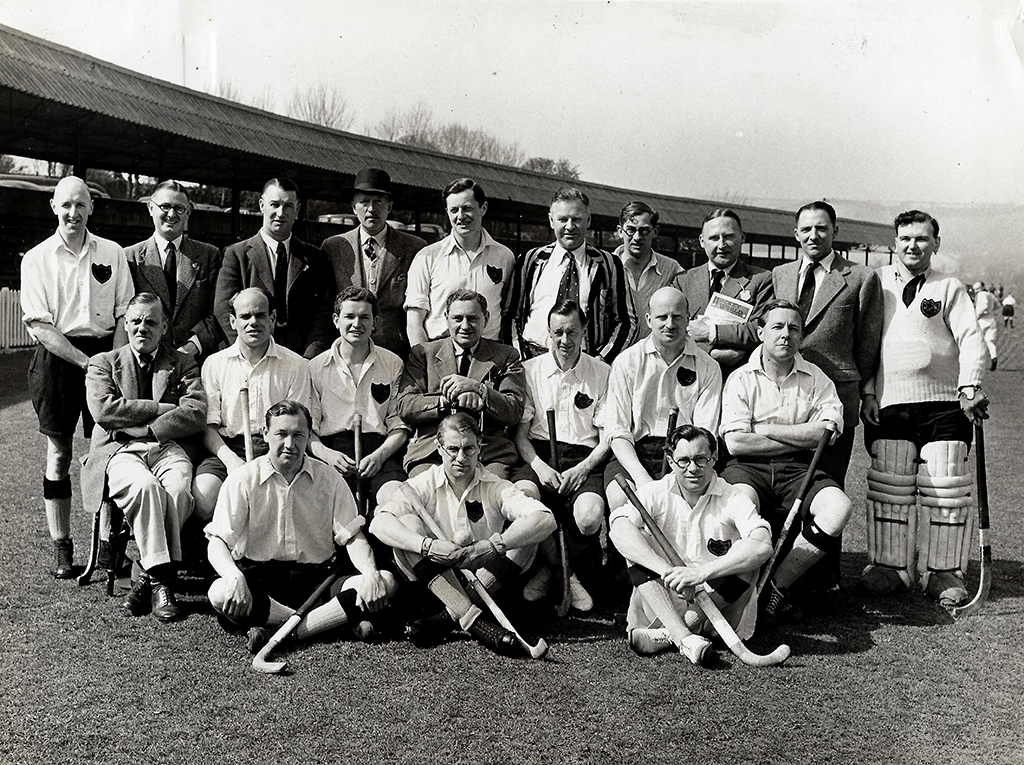 Black and white group photograph of two men's hockey teams standing or seated in three rows. Players are wearing buttoned-up shirts or blazers. They are squinting into the sunshine.