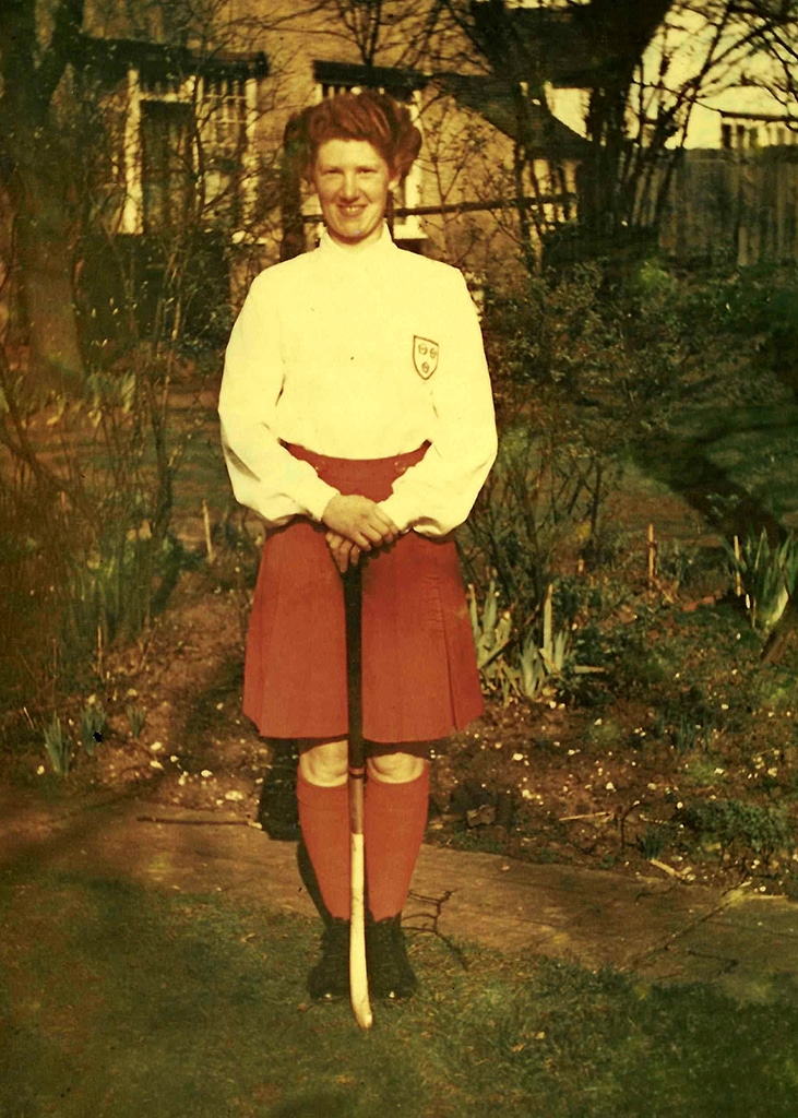 A smiling young lady with auburn hair and wearing a white blouse, red skirt and red socks. She is holding a hockey stick and standing on a lawn in front of a sparse flower bed.