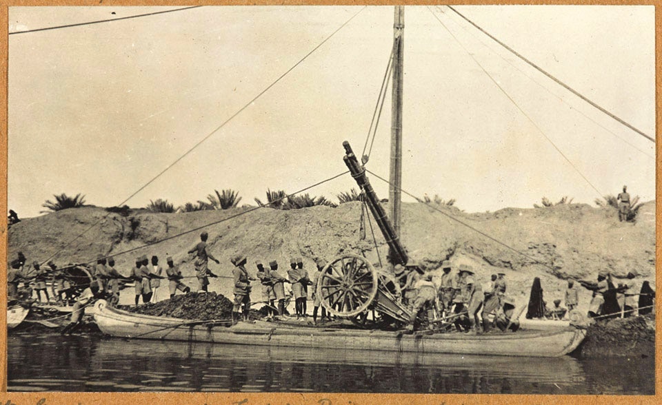 A huge wheeled gun in being hoisted onto a long, narrow boat. People is desert uniforms pull on long ropes to lift and stabilise the gun.