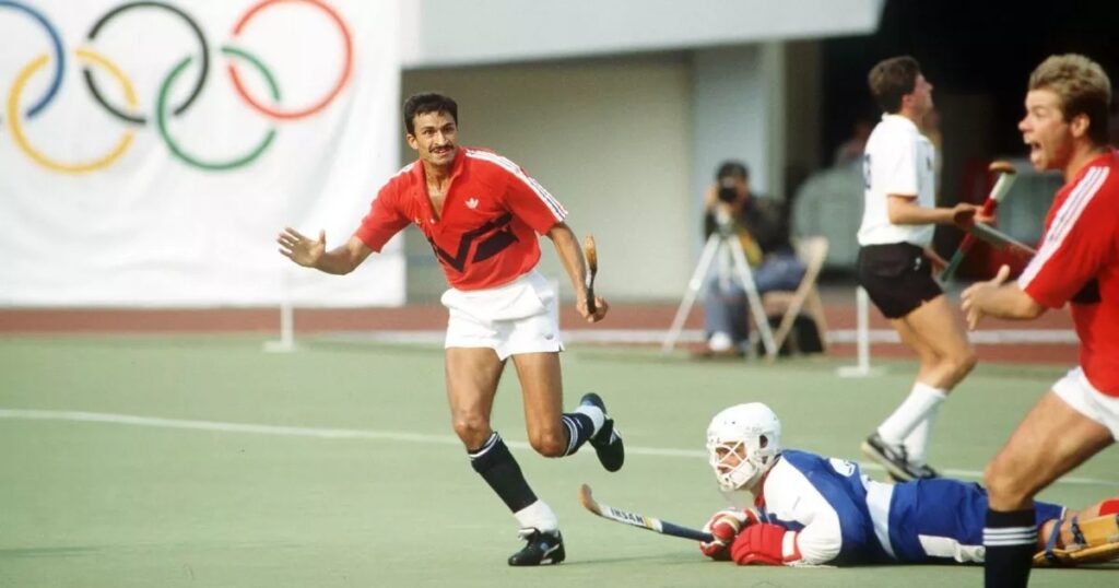 A man of Asian heritage with short, dark brown hair and a moustache wheels away after scoring with one arm held up in celebration. A goalkeeper lies prone on the floor.