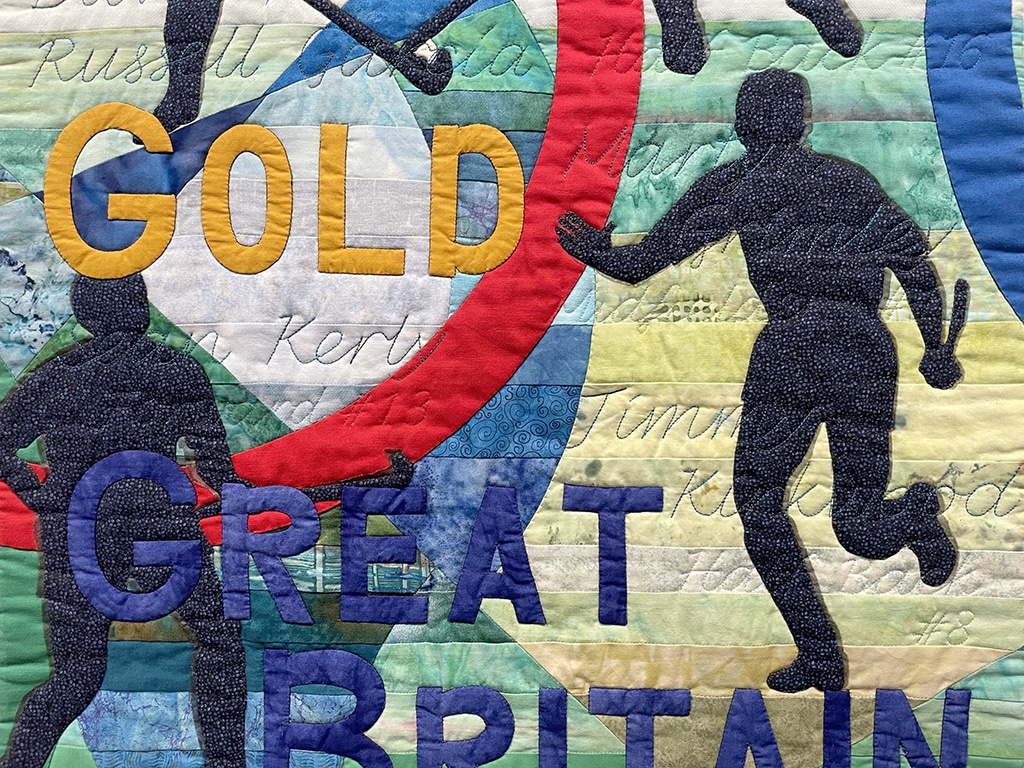 A multi-coloured quilt made up of various fabrics. The word "GOLD" is alongside a silhouette of a man running with one arm held aloft in celebration.