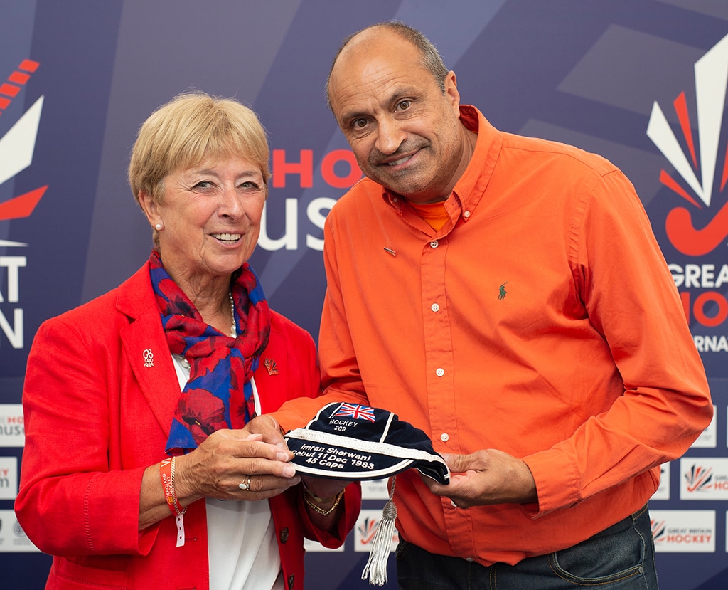 A Caucasian woman presents a navy blue cap to a tall man of South Asian heritage.