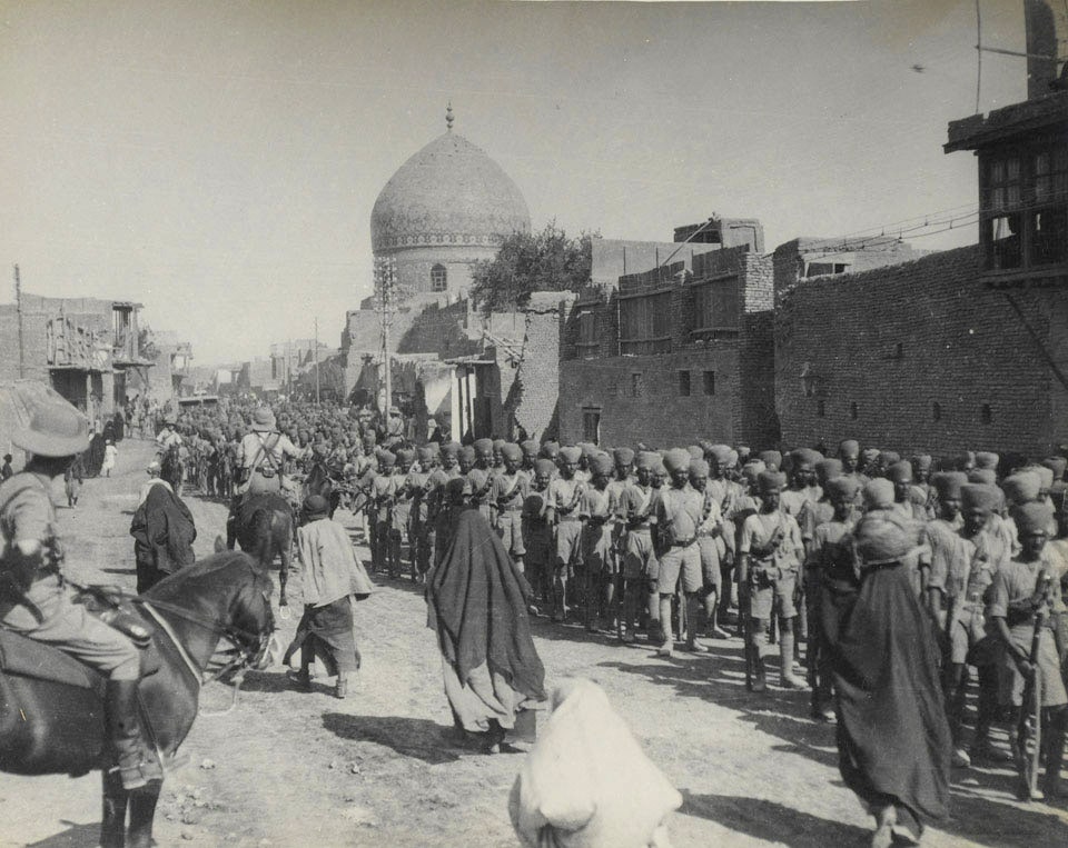 Black and white photograph of a procession of Indian soldiers in turbans marching through a middle Eastern city. Local women walk past in the opposite direction.