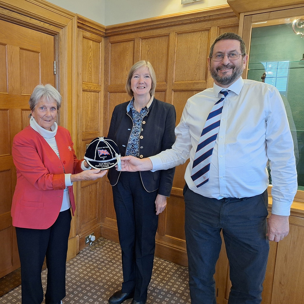 A man and a woman present a navy blue cap to another woman in a room with wood-panelled walls.