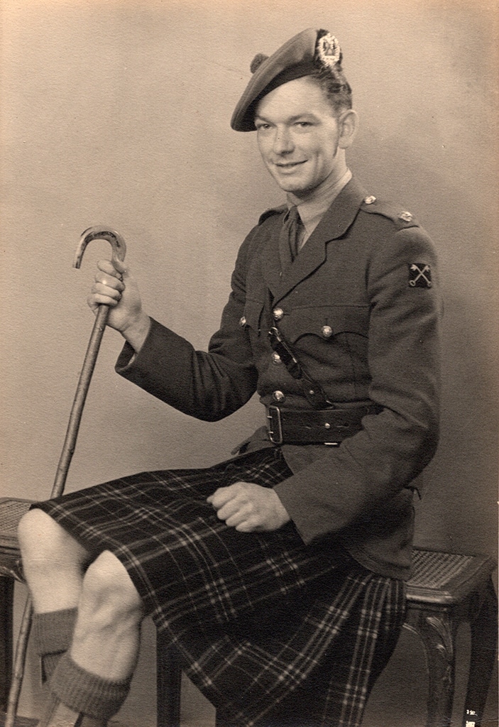 A seated man wearing Scottish miliary uniform including a scotch bonnet and tartan kilt.